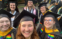 A group of smiling friends in Commencement attire and colorful stoles pose for a selfie