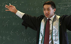 A student in Commencement attire gesturing to a class with a chalkboard full of math equations behind him