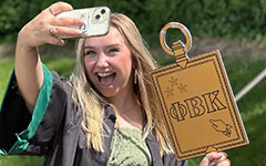 A smiling graduate takes a selfie holding a Phi Beta Kappa sign