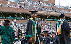 A student smiles and points at the camera as they walk to their seat on the field of Zable Stadium during the Commencement ceremony