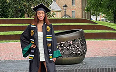 A smiling graduate poses in Commencement attire by the hearth of the Memorial to the Enslaved on historic campus
