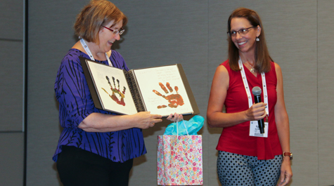 Heather Macdonald stands on stage and is presented with a book of SAGE 2YC Change Agent hand prints