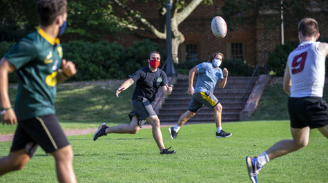 Students wearing masks run down a grassy field, chasing a rugby ball