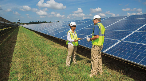Two people in hard hats stand near solar panels in a large field