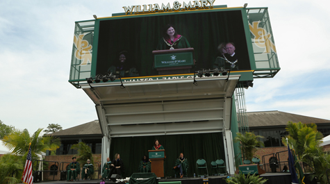 A large screen shows a person at a podium and below is that person on a stage