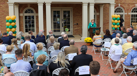 People sit in folding chairs facing a person at a podium in front of a brick building