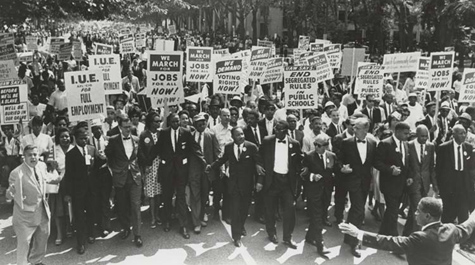 A black and white photo shows a large group of people marching and holding signs about employment
