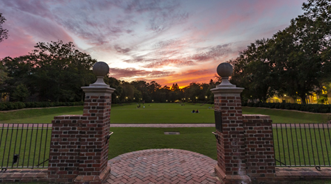 An orange and purple sunset is visible beyond two brick pillars in front of a large green space