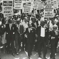 A black and white photo shows a large group of people marching and holding signs about employment