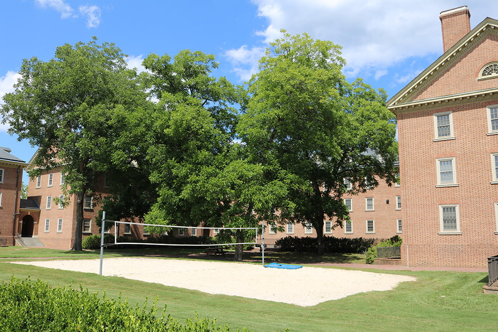 A sand volleyball court surrounded by greenery and a brick, windowed dorm building.