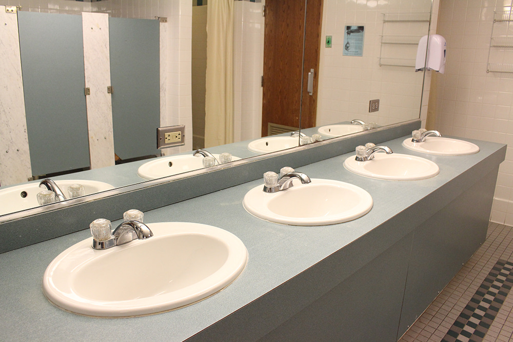 A bathroom with a four-sink vanity reflects toilet stalls and a shower in the mirror.