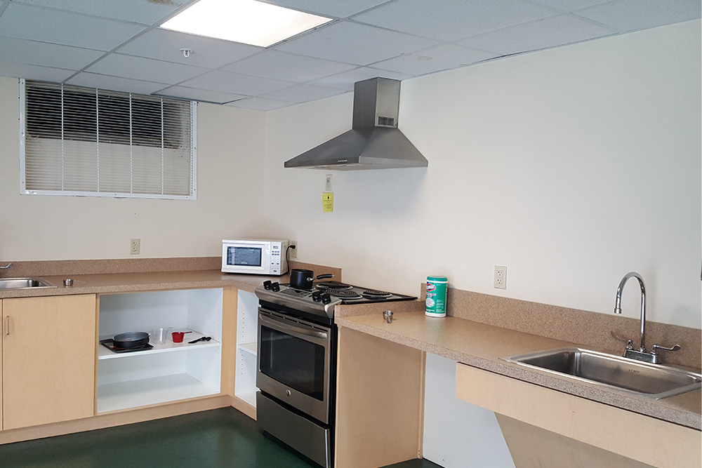 Dorm kitchenette with two sinks, microwave, oven, range hood, and smaller kitchen items on a lower shelf. 