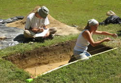 Once excavation is completed, measured drawings are made of the soil strata visible in the test unit wall. One archaeologist reads soil depths to her partner, who sketches the profile on graph paper.