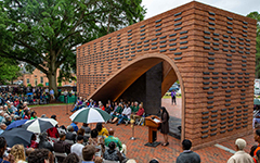 People sitting in chairs surround a brick structure where someone is speaking at a podium