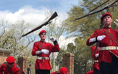 The Queens' Guard, decked in red and black uniform, present their rifle drill in front of a brick fence. 