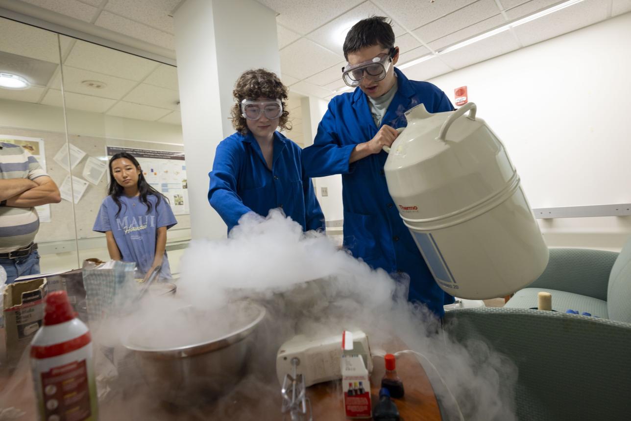 Two students conducting chemistry experiment with fog forming in the foreground and two people onlooking