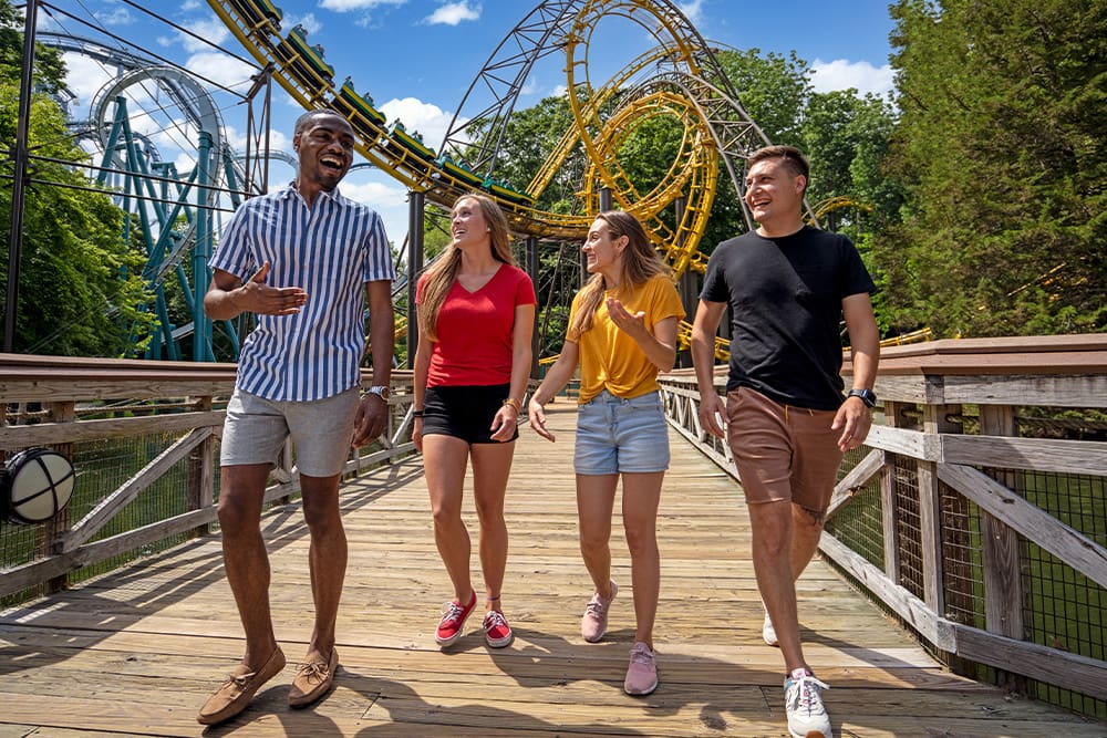 Four smiling people crossing a wooden bridge in the Busch Gardens Williamsburg theme park, with roller coasters in the background