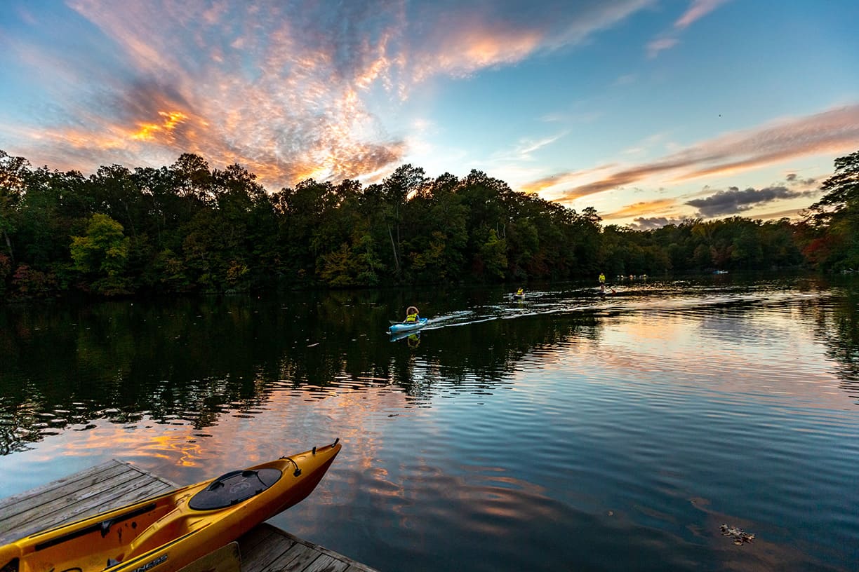 Sunset over a tree-lined Lake Matoaka with people kayaking and stand-up paddle boarding on the water