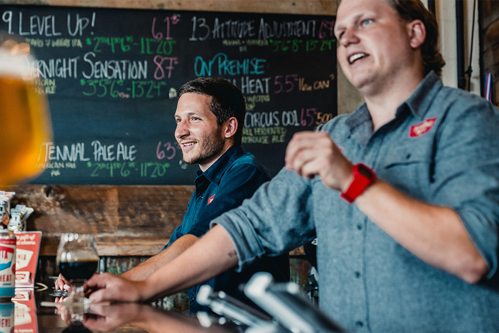 Two Virginia Beer Company employees talking to customers at the bar with a chalk board of craft beer selections in the background