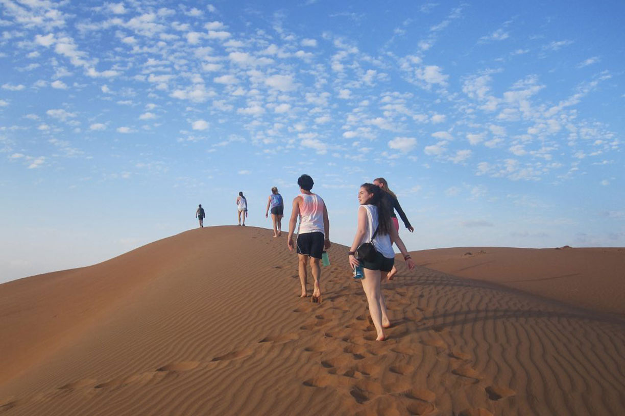 Six students walk up a sand dune with blue skies above
