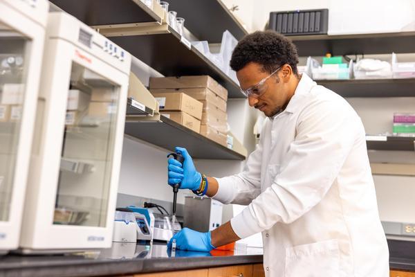 A student in a lab coat, blue gloves and goggles fills a test tube in a science lab