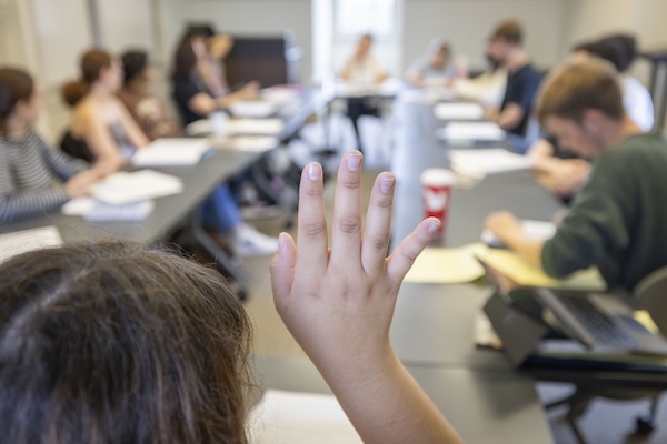 A student raises their hand in the foreground of a roundtable class discussion
