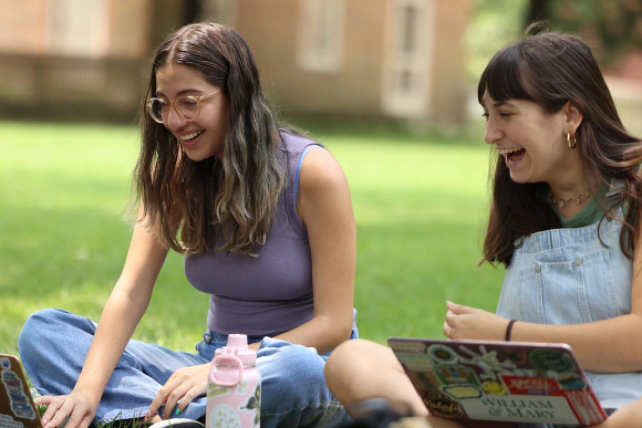 Two students sit on the grass with their laptops