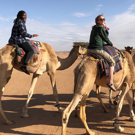 Two smiling students riding camels in the desert of Oman on a geology study abroad trip.