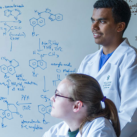 Two students in lab coats in front of a whiteboard of chemistry diagrams