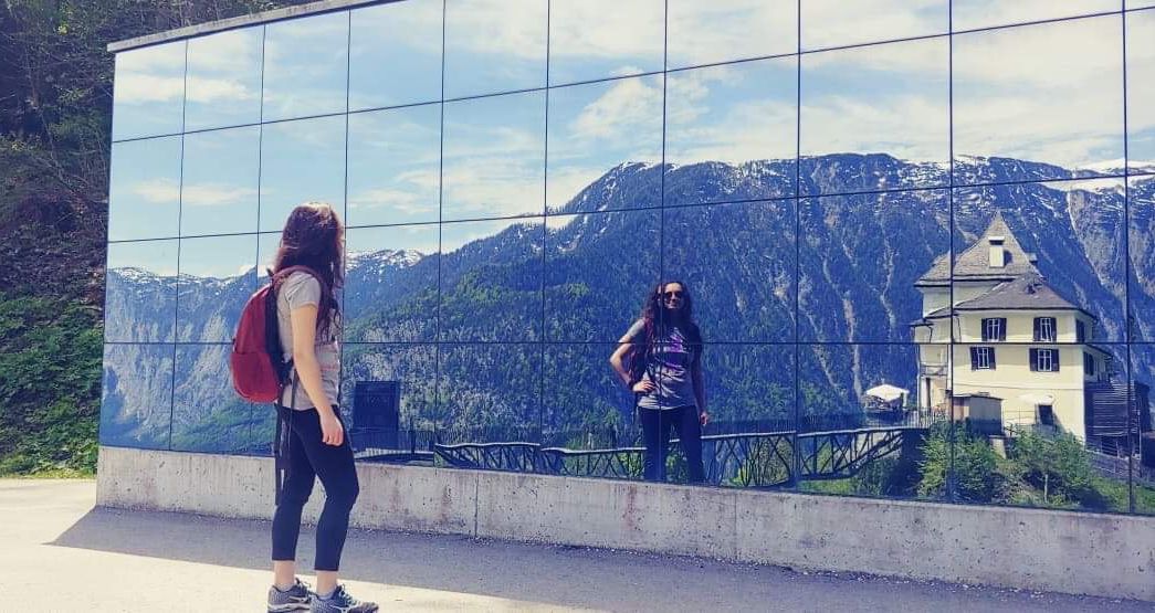 Gabriella Carney ('18) standing with her back to the camera in front of a mirror-reflective glass building. The reflection shows Gabriella with her hand on her hip with a mountain in the distance.