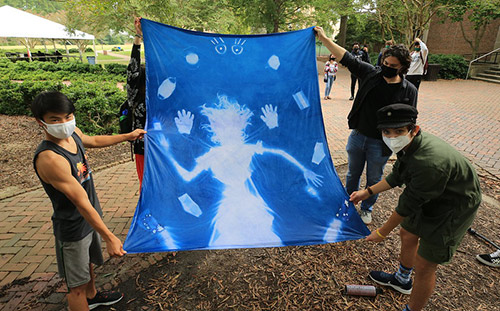 Masked students inspect the cloth mural panel they created in "Photomania" class where they worked on cyanotypes
