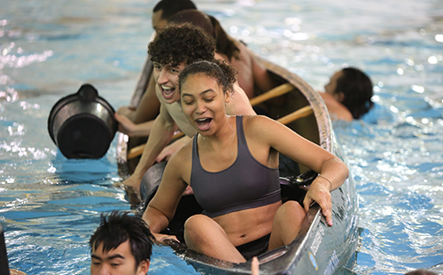 Students laughing in a tipping canoe in an indoor swimming pool filling buckets of water.