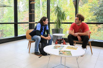 Two students seated in conversation in the atrium of the McLeod Tyler Wellness center with a serene view of the woods through the wall of windows behind them.