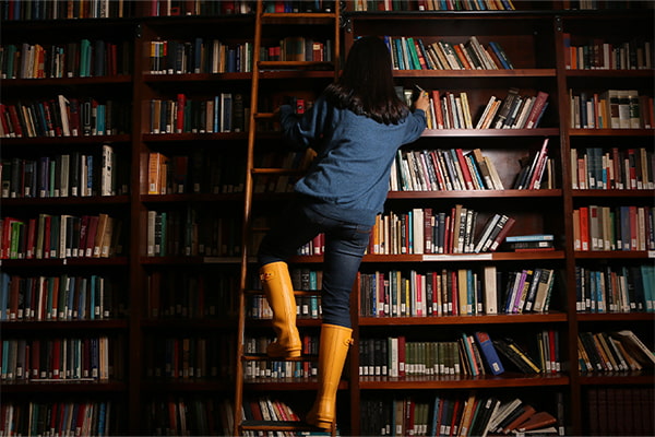 Tall shelves of books in a library with a student on a rolling ladder