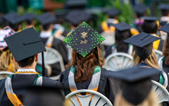 Graduates in caps and gowns sit facing away from the camera, and one cap is decorated with a W&M cypher and 2021