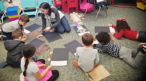 A teacher sits on the ground with several children who are working on an art project