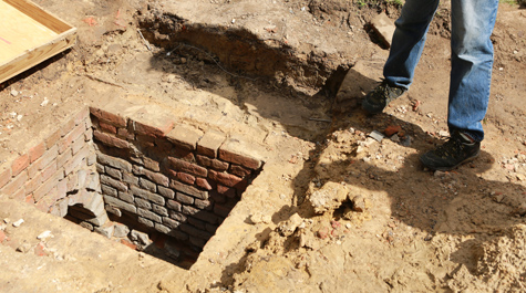 Someone's feet are visible near a hole in the ground that reveals a vaulted brick drain