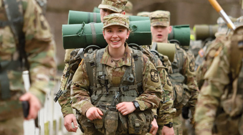 A soldier in uniform smiles at the camera while marching with others