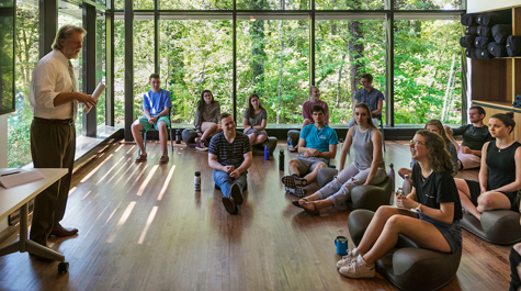 Students sit on the floor of a room surrounded by glass windows and listen to Kelly Crace