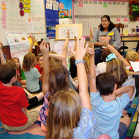 Elementary school children raise their hands while facing a teacher sitting on a chair