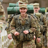 A soldier in uniform smiles at the camera while marching with others