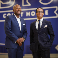 Two men smiling and wearing suits stand in front of a blue background