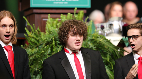 Members of Phi Mu Alpha Sinfonia dressed in black coats, white shirts and red ties, singing