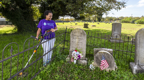 A person stands next to a headstone and holds a weedwhacker