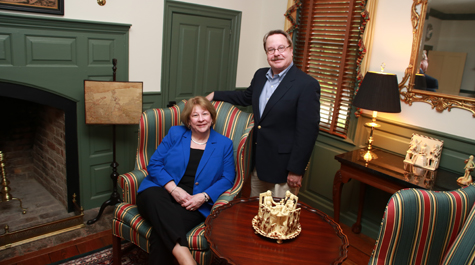 One person sits in a chair and another person stands by the chair inside a living room