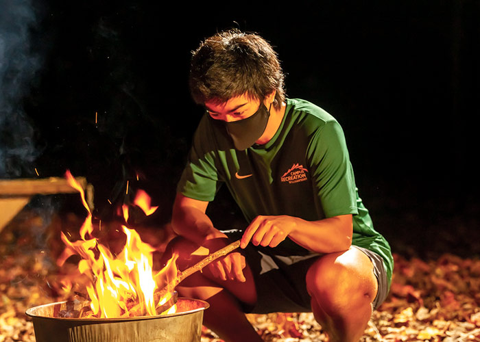 A masked student in a Campus Rec shirt tends to a fire bowl at night in a wooded setting.