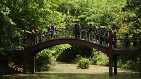 People in academic regalia walk over a curved wooden bridge