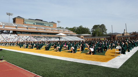 People in caps and gowns sit in chairs on a football field