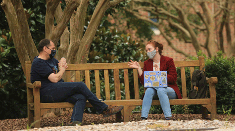 Two people wearing masks talk while sitting on a bench