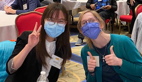 Two people wearing masks gesture with a peace sign and thumbs-up